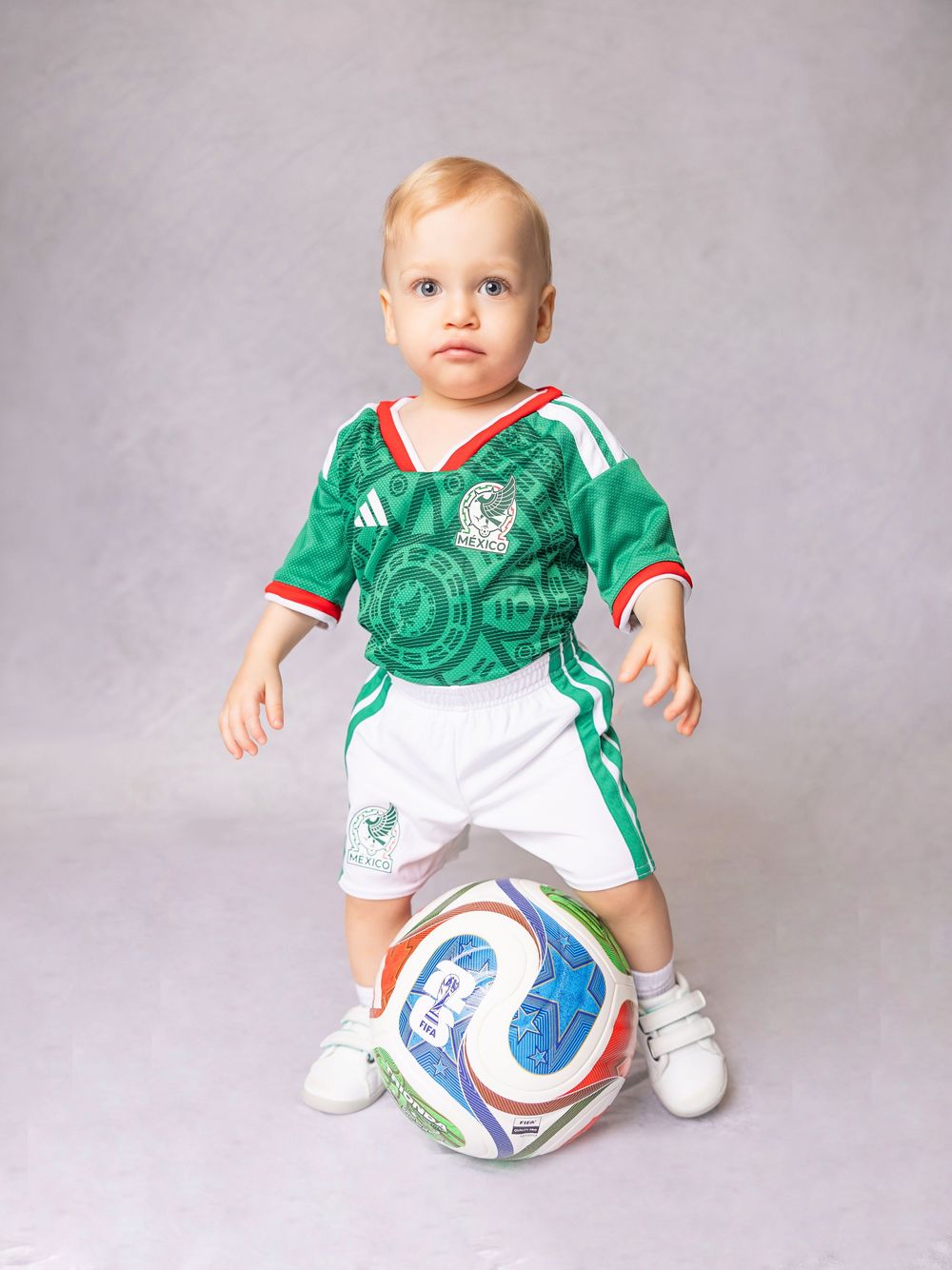 Baby in green Mexico soccer uniform with white shorts and sneakers, standing next to a colorful soccer ball.