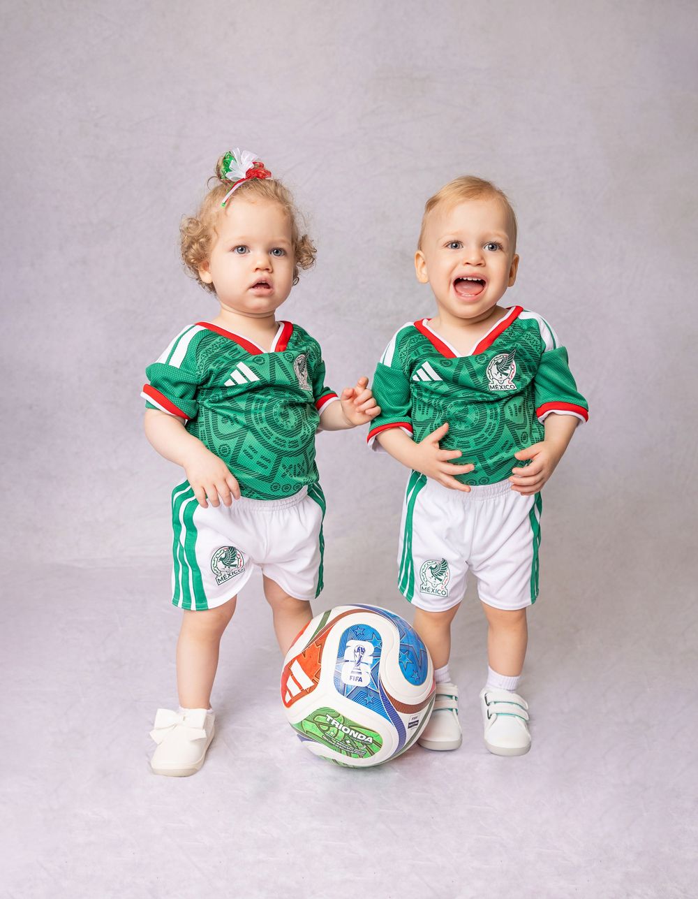 Two toddlers in matching Mexico soccer jerseys and white shorts pose with a colorful soccer ball.