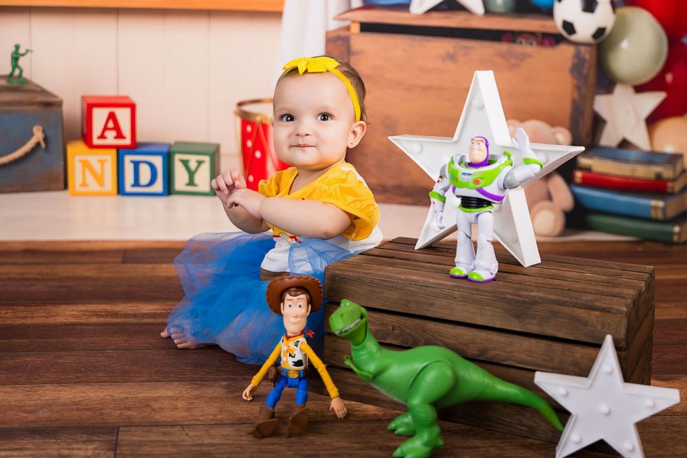 Baby in yellow shirt and blue tutu sits with Toy Story characters Woody, Buzz, and Rex near ANDY blocks.