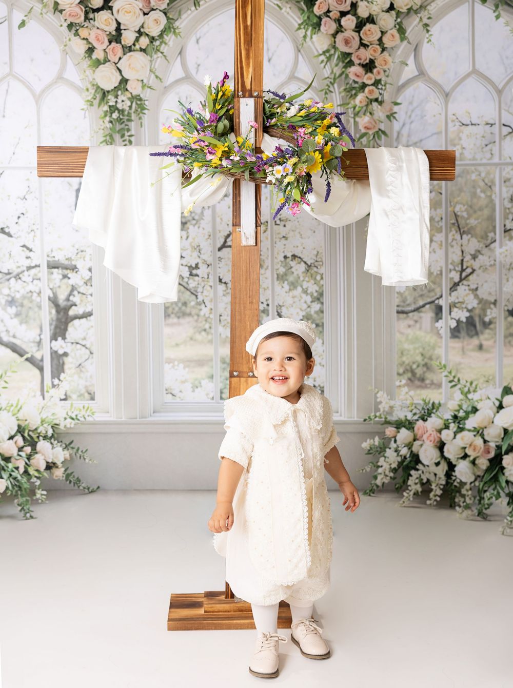 Baby in white christening outfit stands before a floral cross in an elegant baptism photo session.