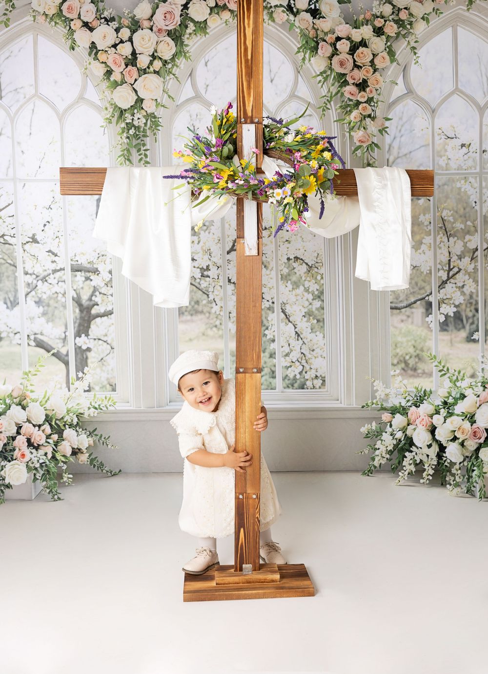 Baby in white outfit holding a wooden cross decorated with flowers in an elegant floral studio setting.