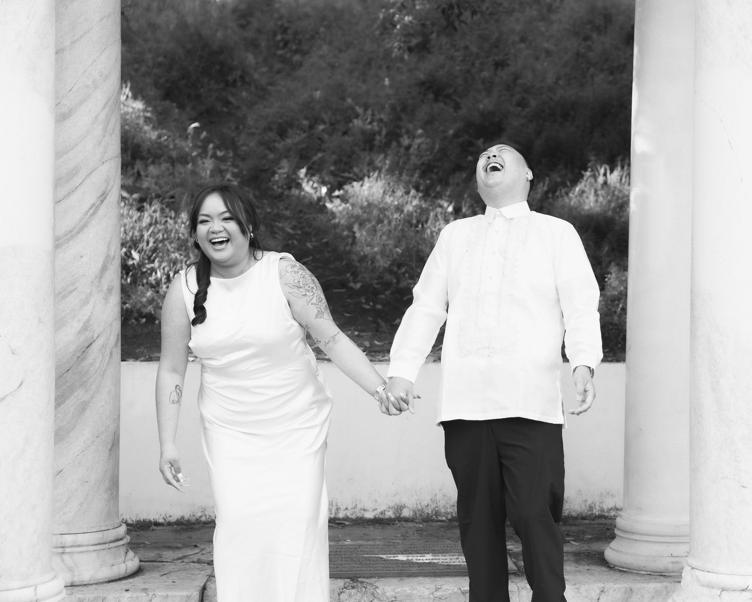A black and white wedding photo of a couple holding hands between white columns.