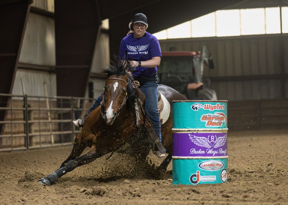 Barrel racer on a brown horse navigating a tight turn around a green barrel in an indoor rodeo arena.