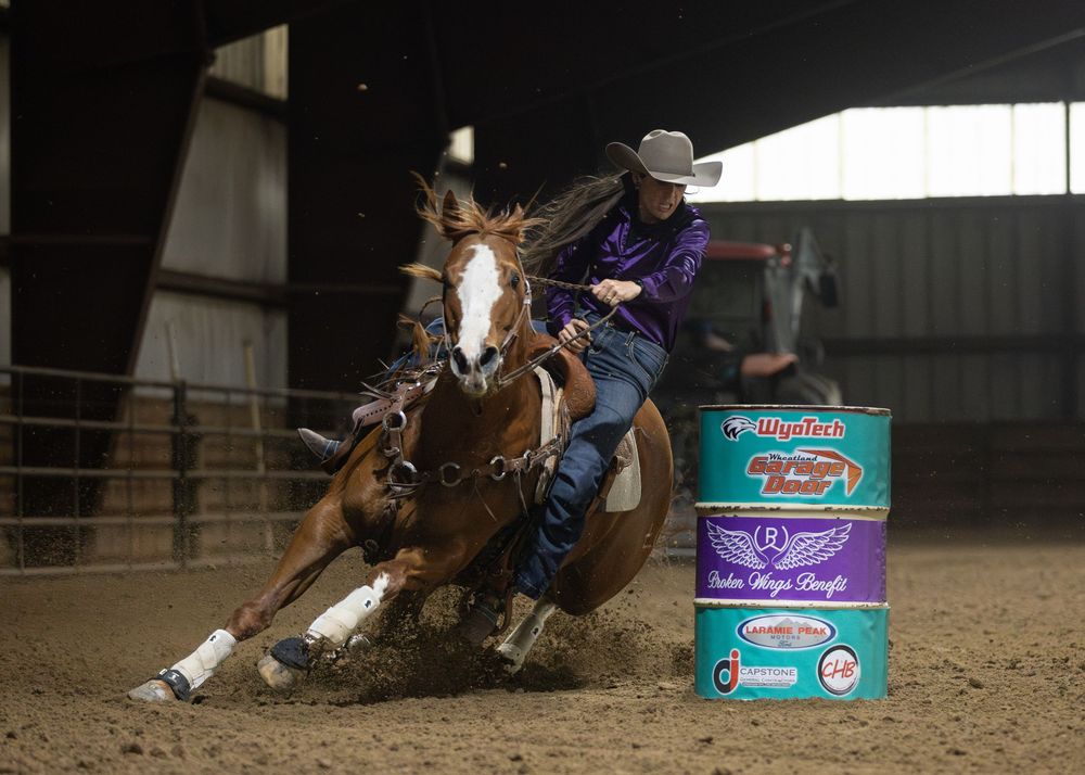 Barrel racer on a chestnut horse navigating a turn around a barrel in an indoor rodeo arena.
