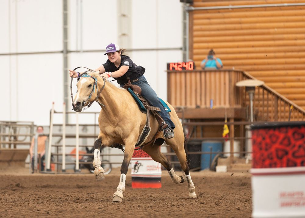 A young female rider on a palomino horse competing in barrel racing at an indoor rodeo arena.