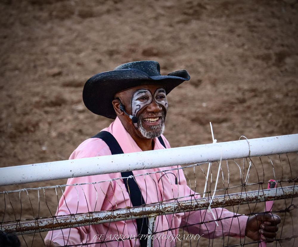 Spectator in pink shirt and cowboy hat smiling while leaning on rodeo arena fence.