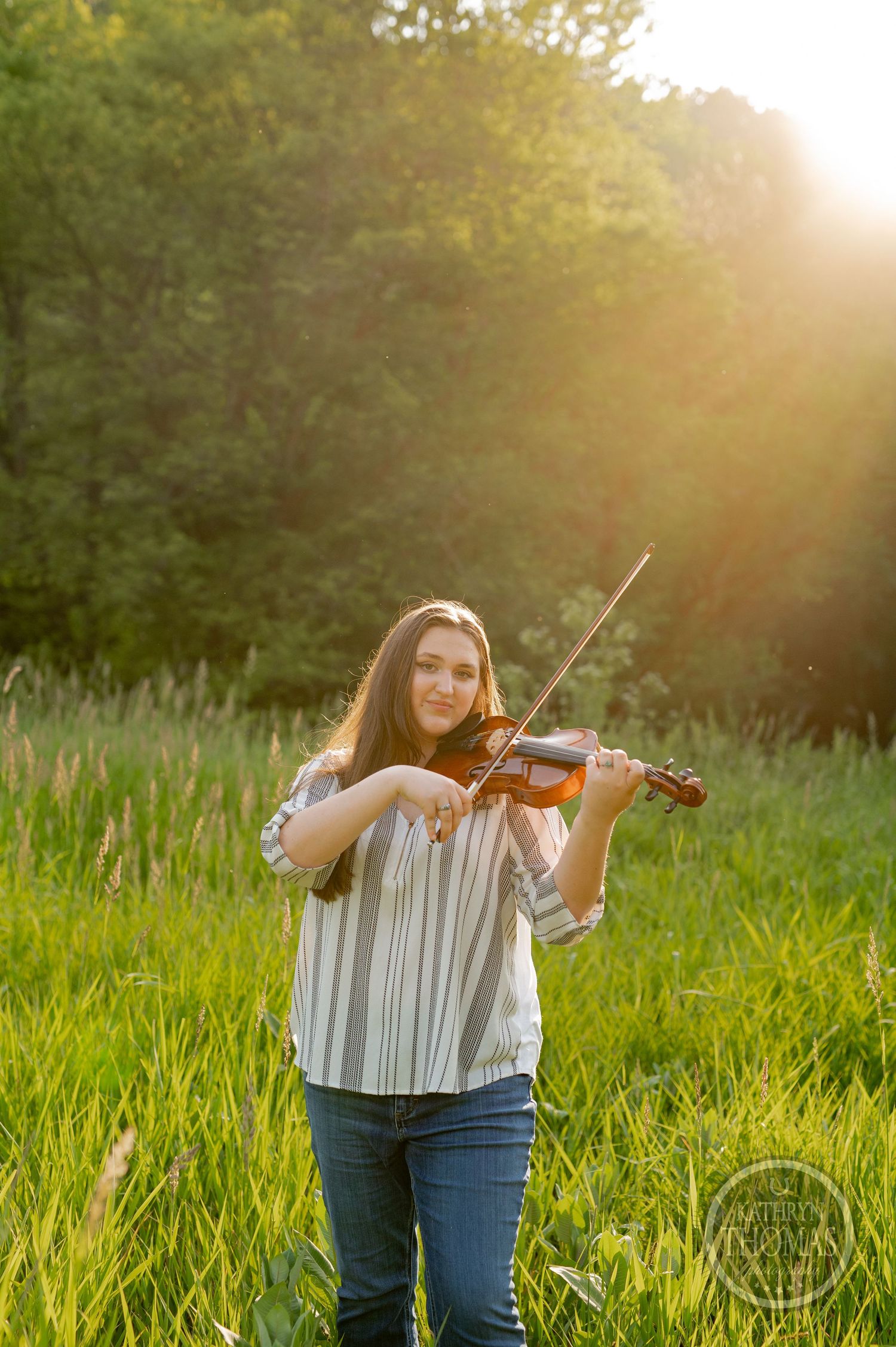 Violinist performs in golden sunlight amidst tall grass and trees.