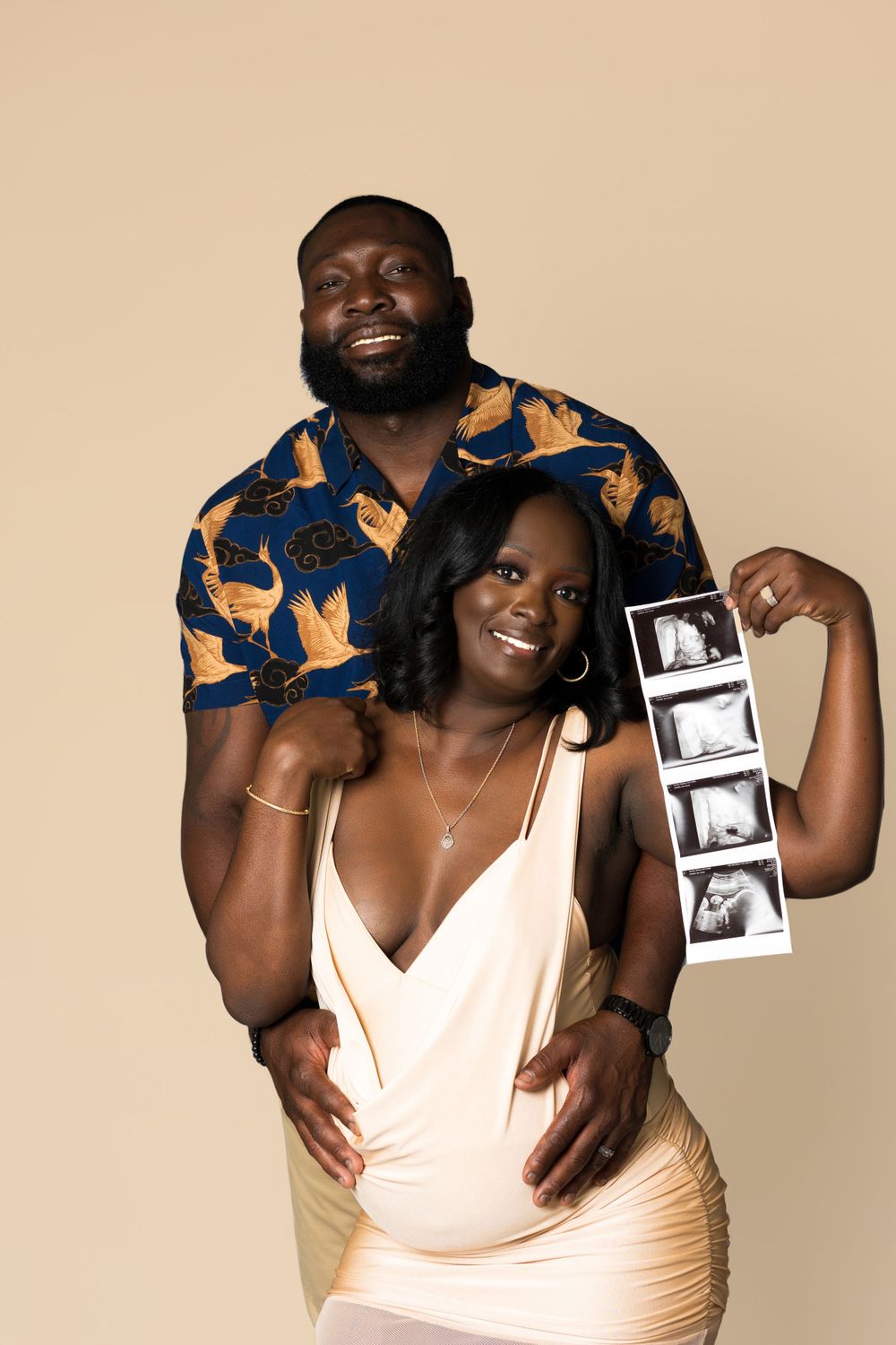 Expectant couple poses with ultrasound photos against beige background.