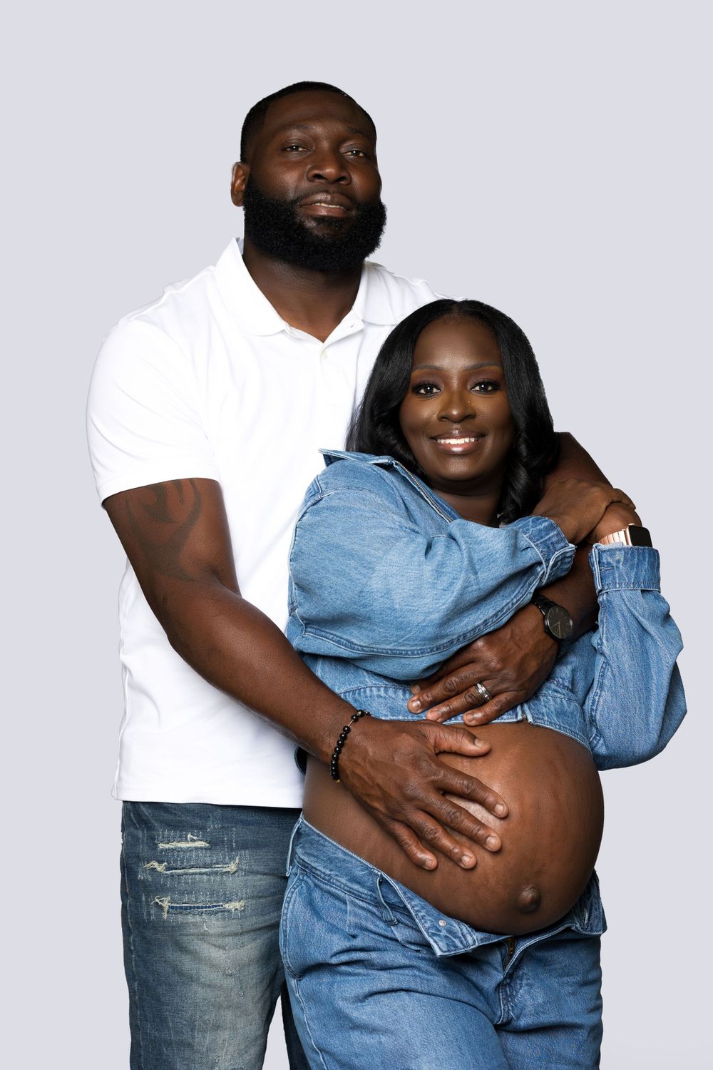 Maternity portrait of couple in casual wear against light background.