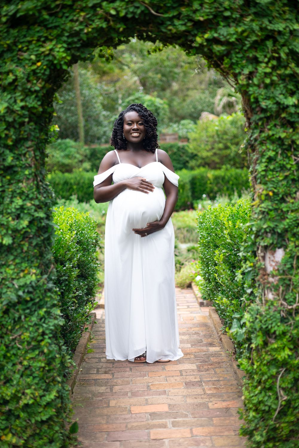 A maternity photo shoot in a garden setting features an elegant white off-shoulder gown on a brick pathway surrounded by greenery.