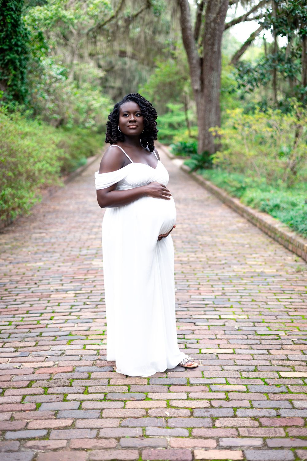 An expectant mother poses in a flowing white dress on a brick path lined with trees and Spanish moss.