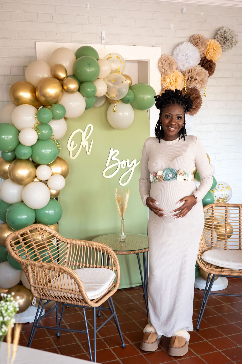 A baby shower setup with green and gold balloon arch, rattan chair, and expectant mother in a cream dress.
