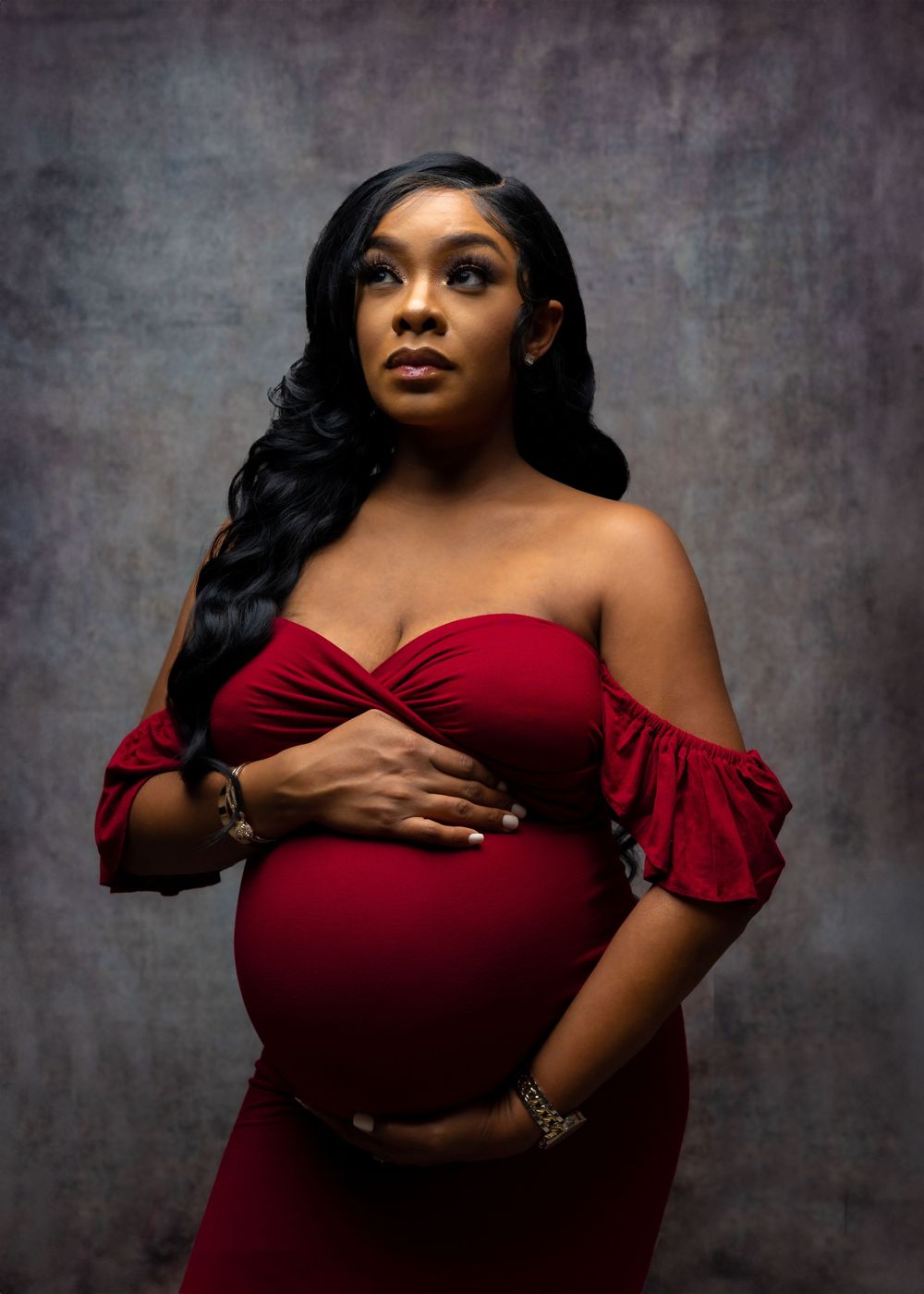 Maternity portrait of woman in red off-shoulder dress against textured gray background.
