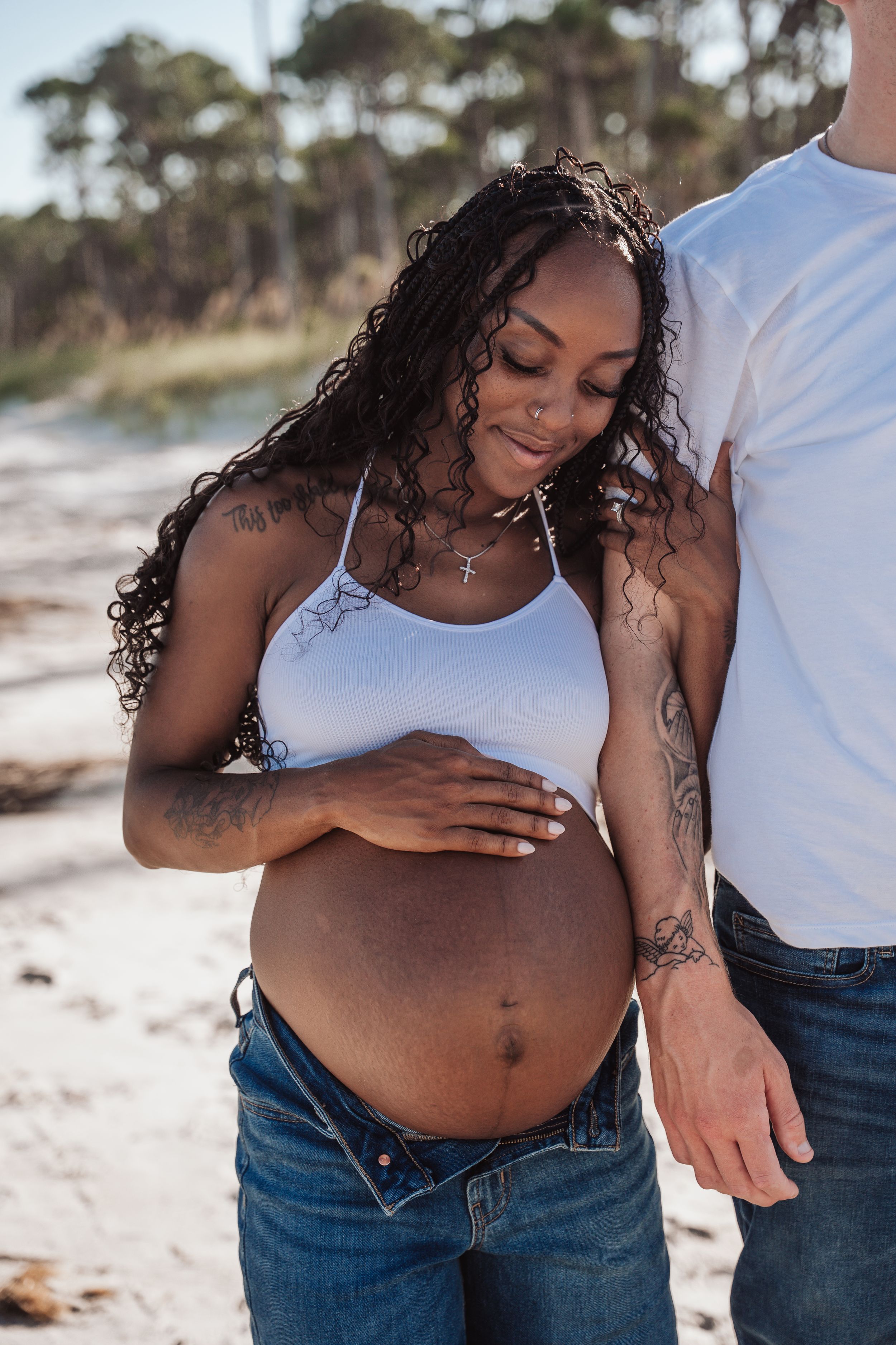 A pregnant woman in a white tank top and jeans stands on a beach, lovingly touching her belly with sunlit trees behind her.