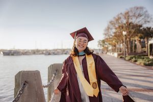 Graduate in maroon cap and gown poses with diploma on sunny campus walkway during graduation celebration.