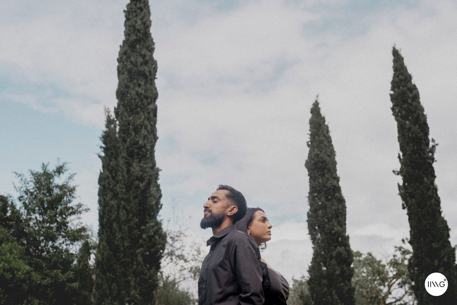 Two people stand close together against a backdrop of tall cypress trees and cloudy sky.