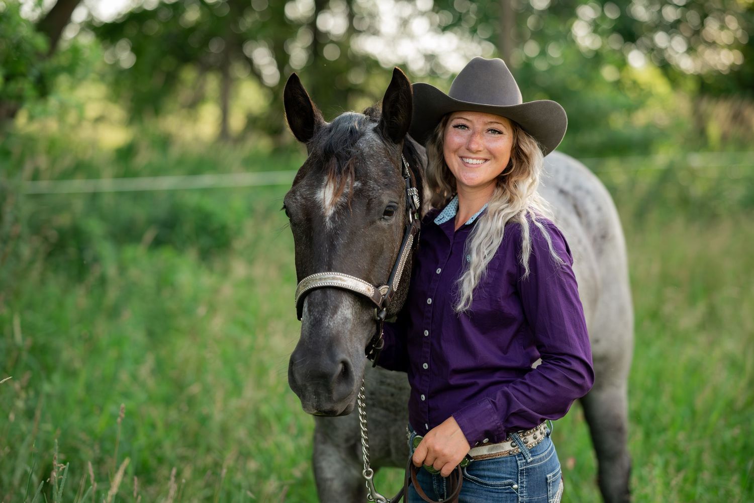 Woman in purple shirt and cowboy hat smiling while posing with black horse in grassy field.