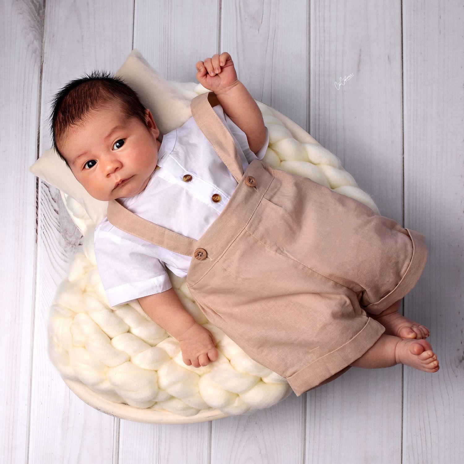 Newborn portrait of baby boy in beige overalls and white shirt laying on white wood bowl on white wood floor looking up at camera.