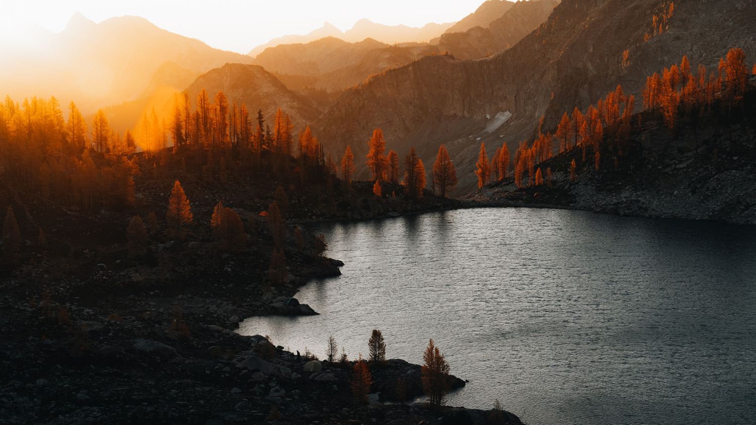 Sunset illuminates the mountain peaks and evergreen trees around a serene alpine lake in vibrant orange and gold hues.