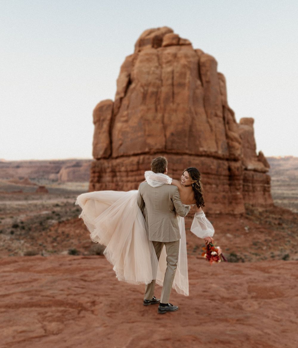 A romantic desert wedding photo with flowing dress against dramatic red rock formations at sunset.