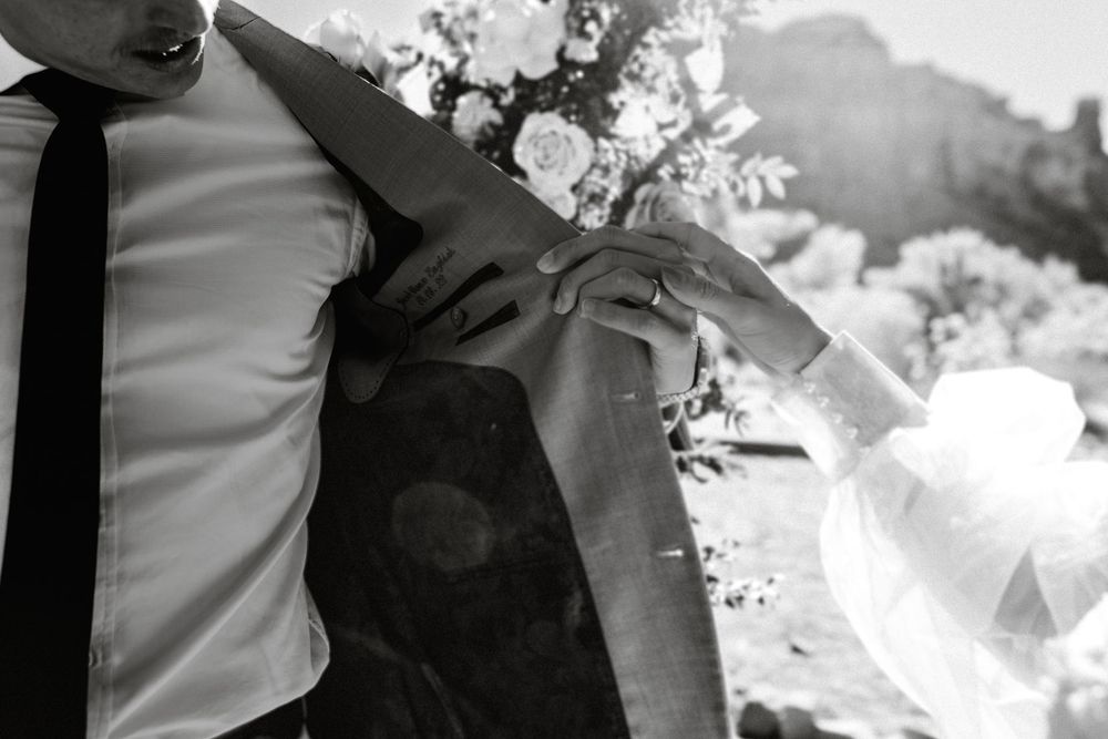 Black and white detail shot shows a groom's suit jacket and tie against a light background.