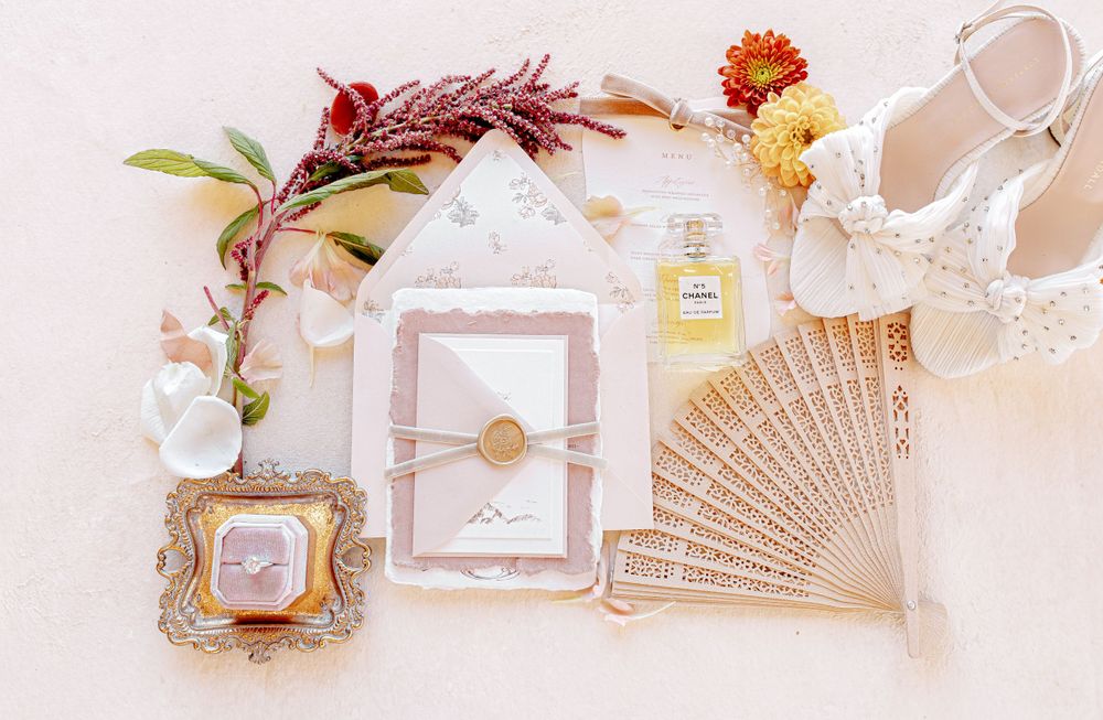 A flat lay of wedding accessories including perfume, shoes, invitations and a decorative fan in coral and pink tones.