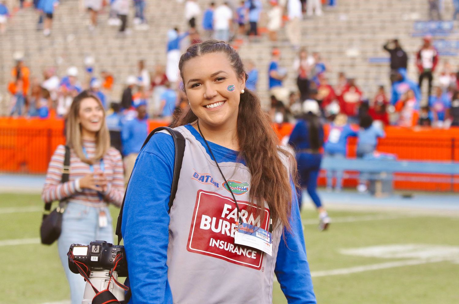Person wearing media credentials smiles on football field with stadium crowd in background.