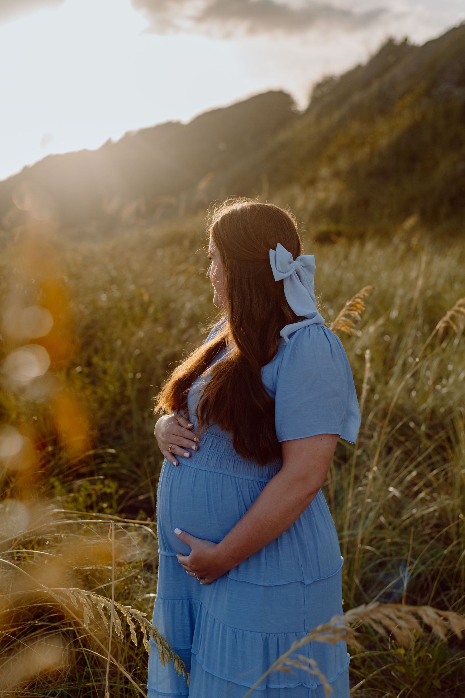 Pregnant woman in blue dress stands in golden field at sunset touching her baby bump.