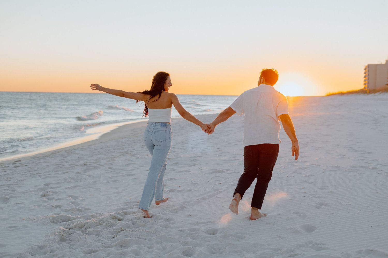 Two people run hand in hand on a beach at sunset.