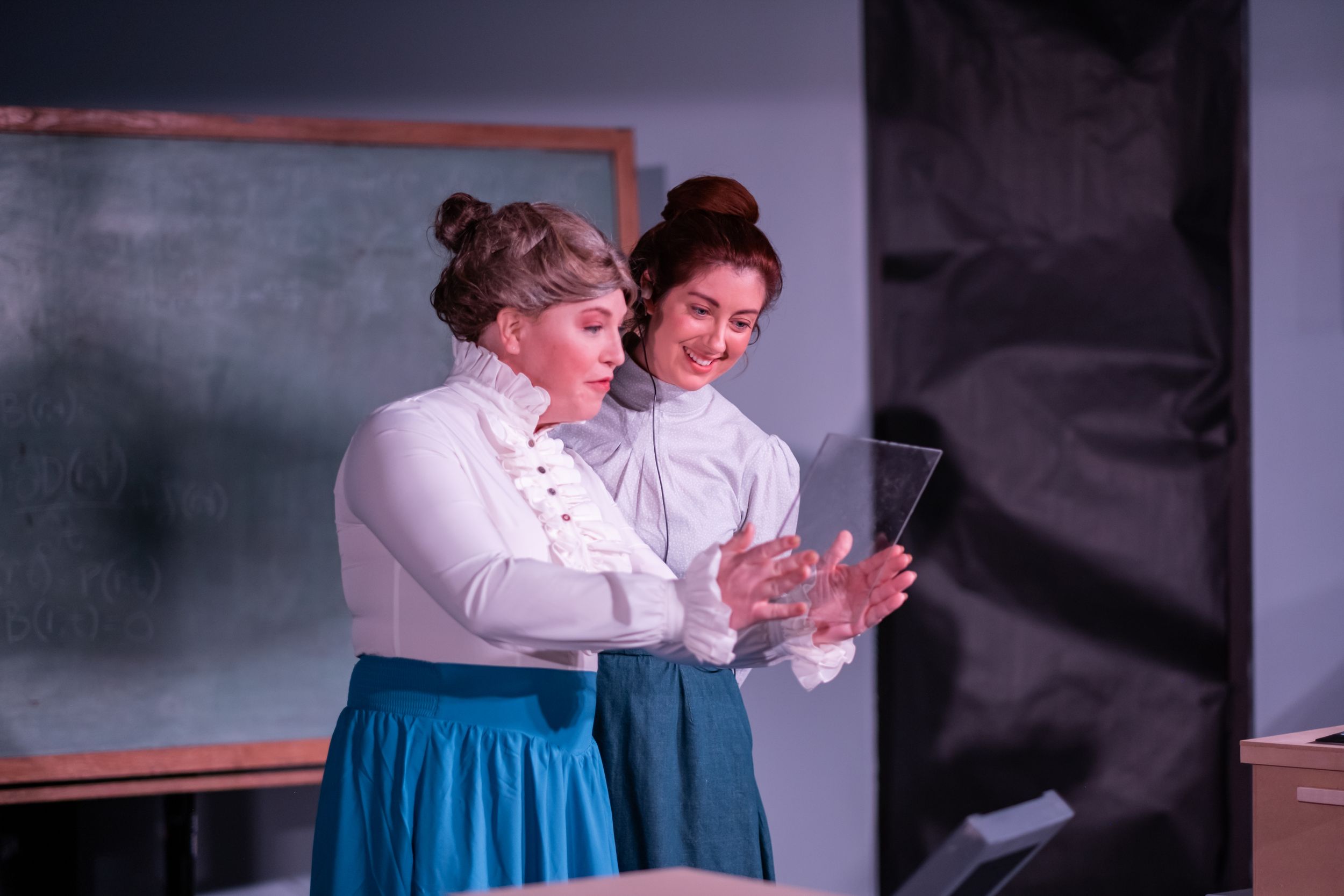 Two performers in period costumes share a moment while looking at something together.