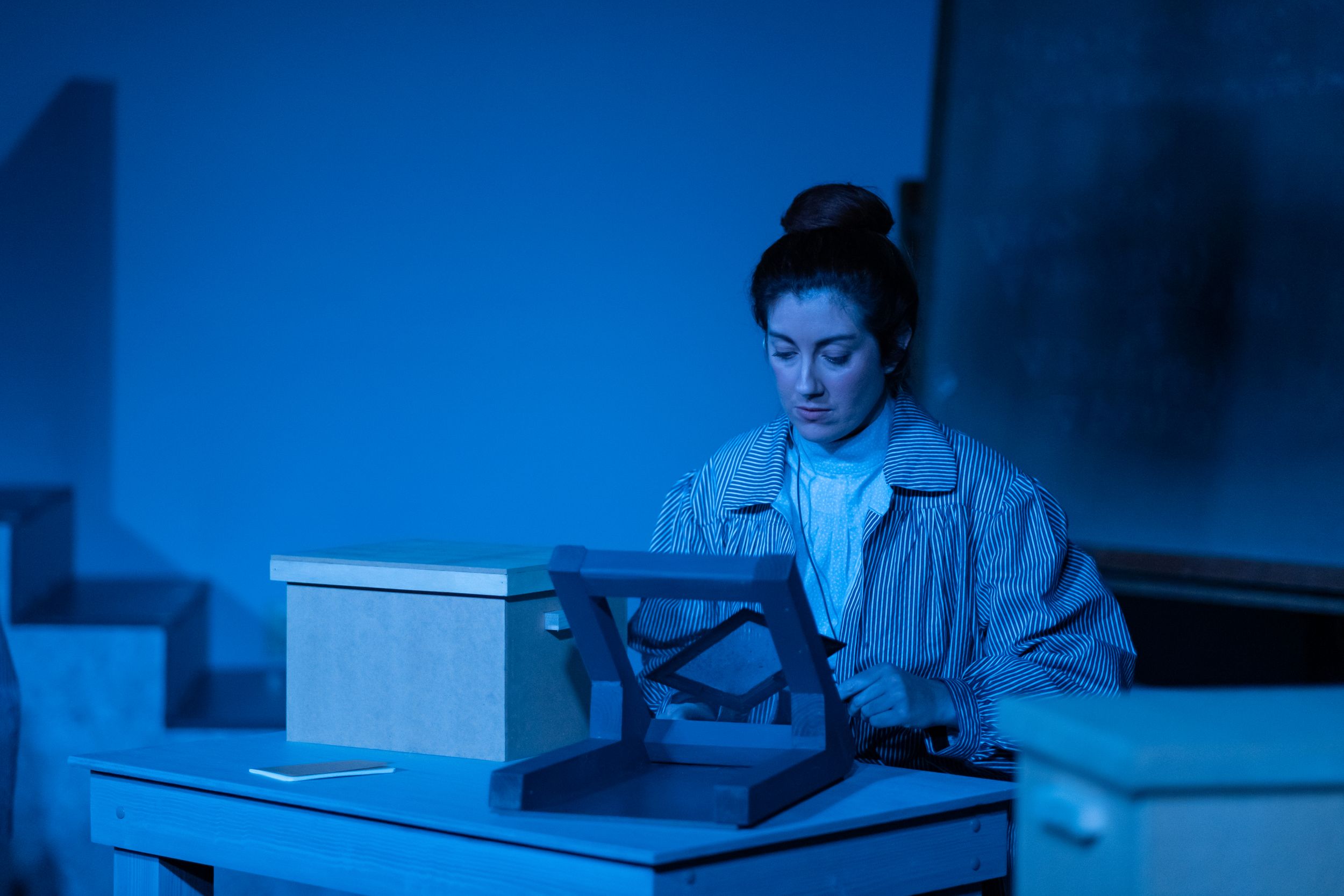 Performer at desk illuminated by blue stage lighting during theatrical scene.