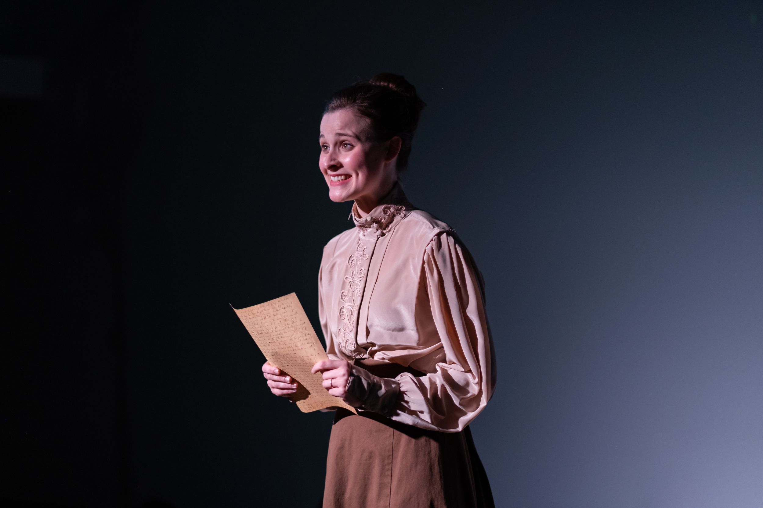 Stage performer in vintage attire reads from paper in dramatic lighting.