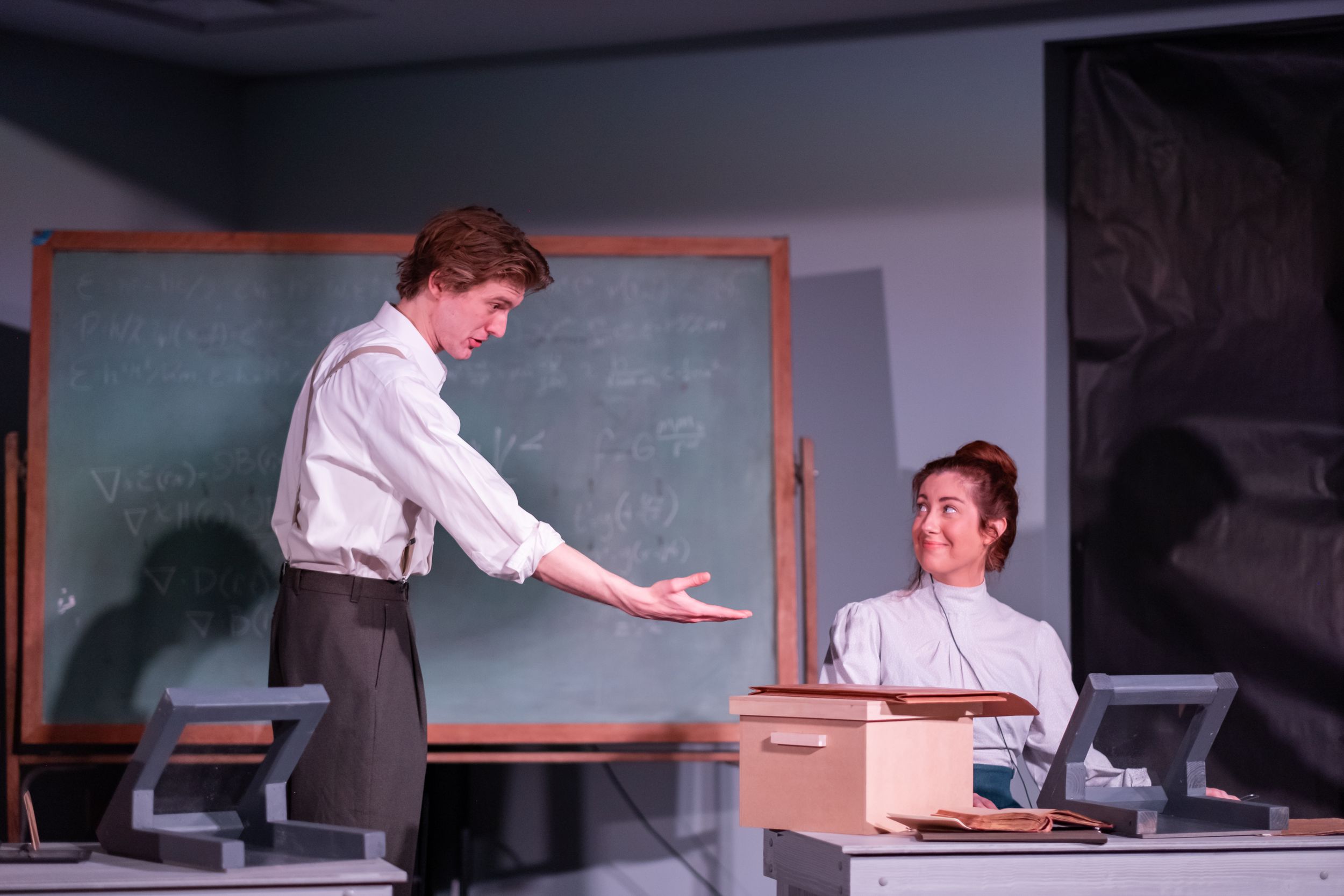 Dramatic scene in a classroom with performers interacting near a chalkboard.