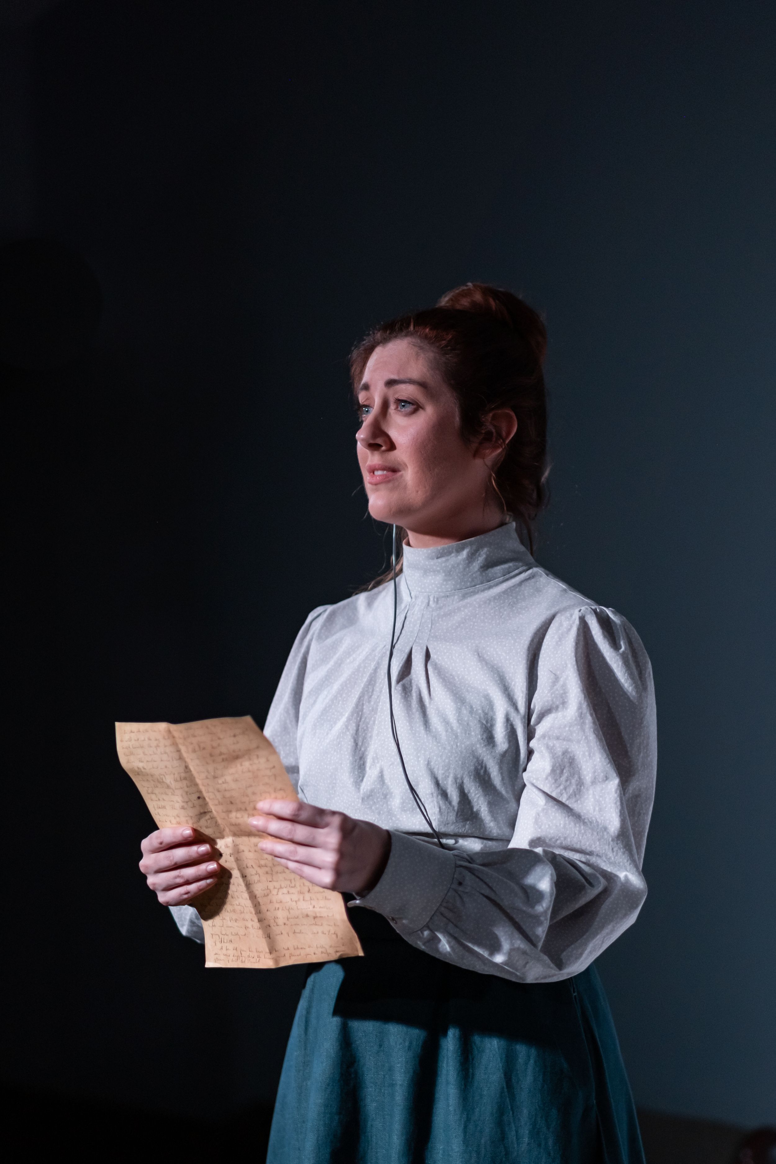 Period drama performer in vintage attire reads from an aged letter against dark backdrop and dramatic lighting.