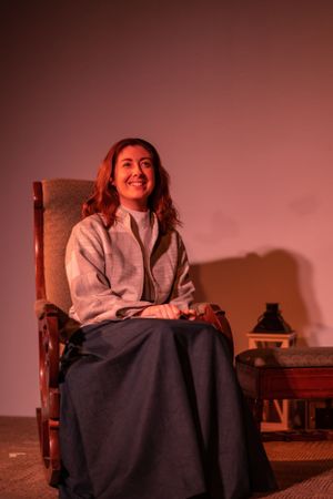 Performance scene of someone in period dress sitting in wooden chair under warm lighting.