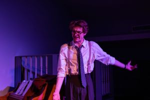 Theater performer in suspenders gesturing dramatically under purple stage lighting.