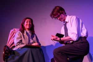Two performers in period costumes engaged in conversation under purple stage lighting.