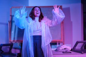 Person in white flowy costume performing dramatically under colorful theatrical lighting.