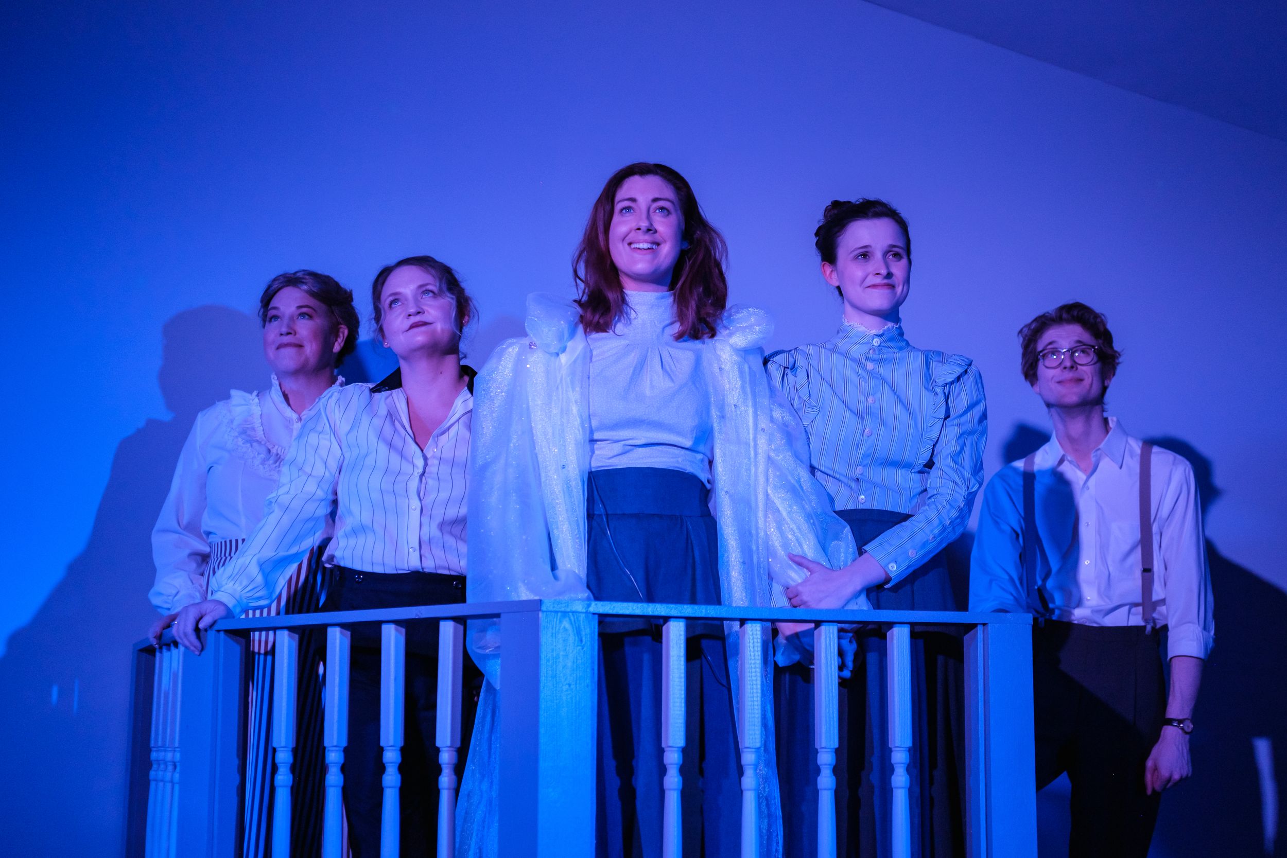 Group posed dramatically at balcony railing under blue theatrical lighting.