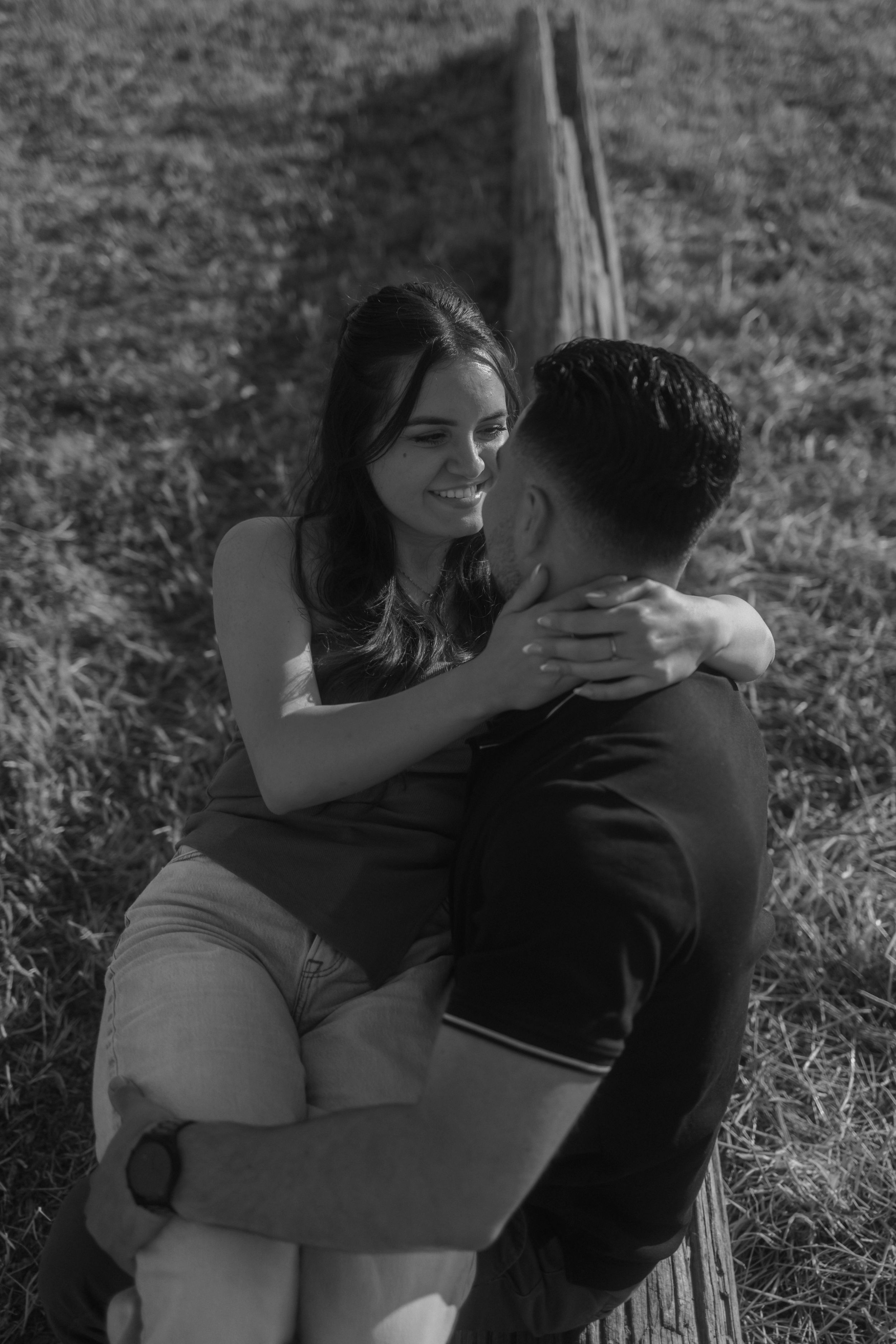 Couple sitting on a grassy field, smiling at each other. The woman is on the man’s lap with her arms around his neck.