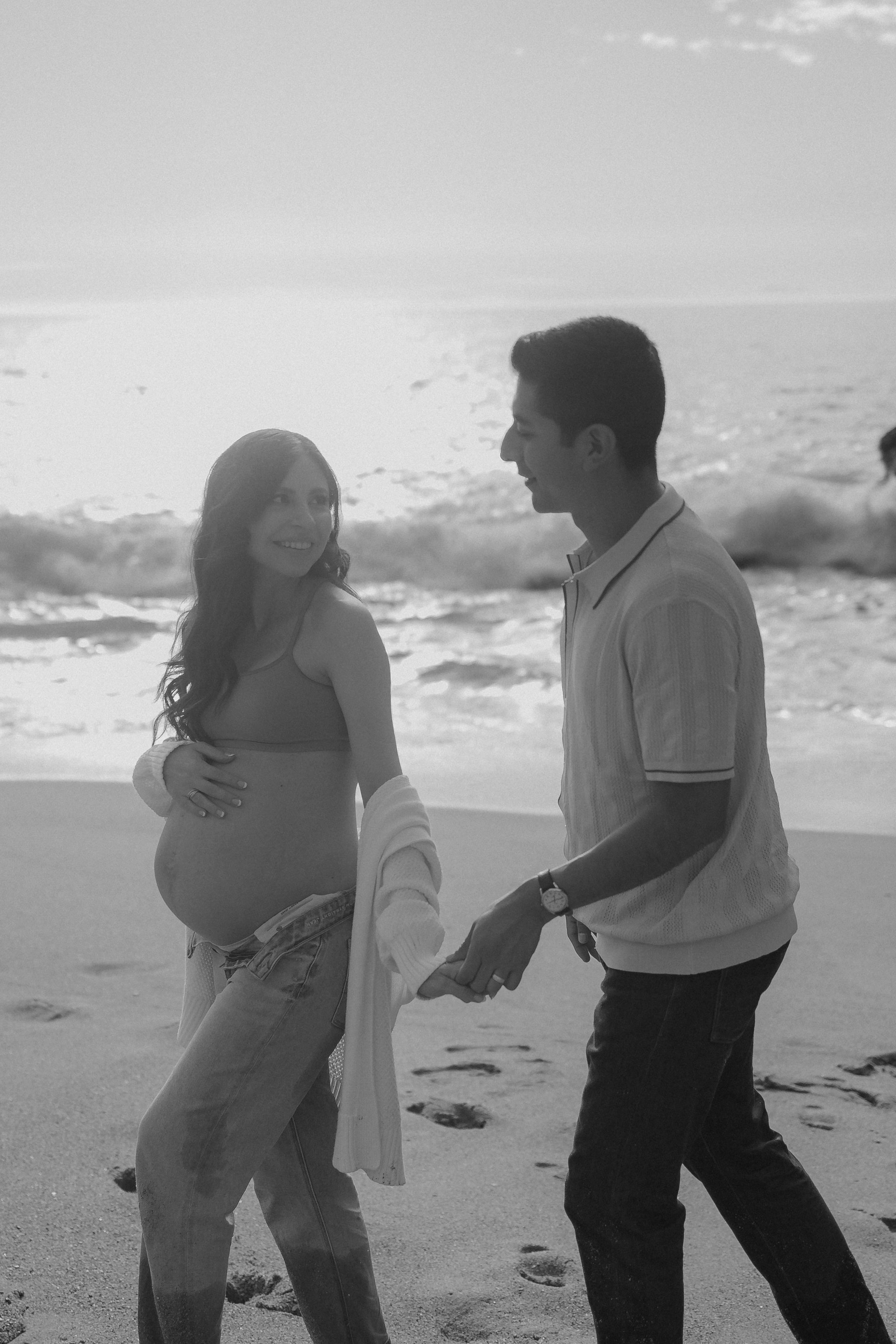 Pregnant woman and man holding hands while walking on a beach.
