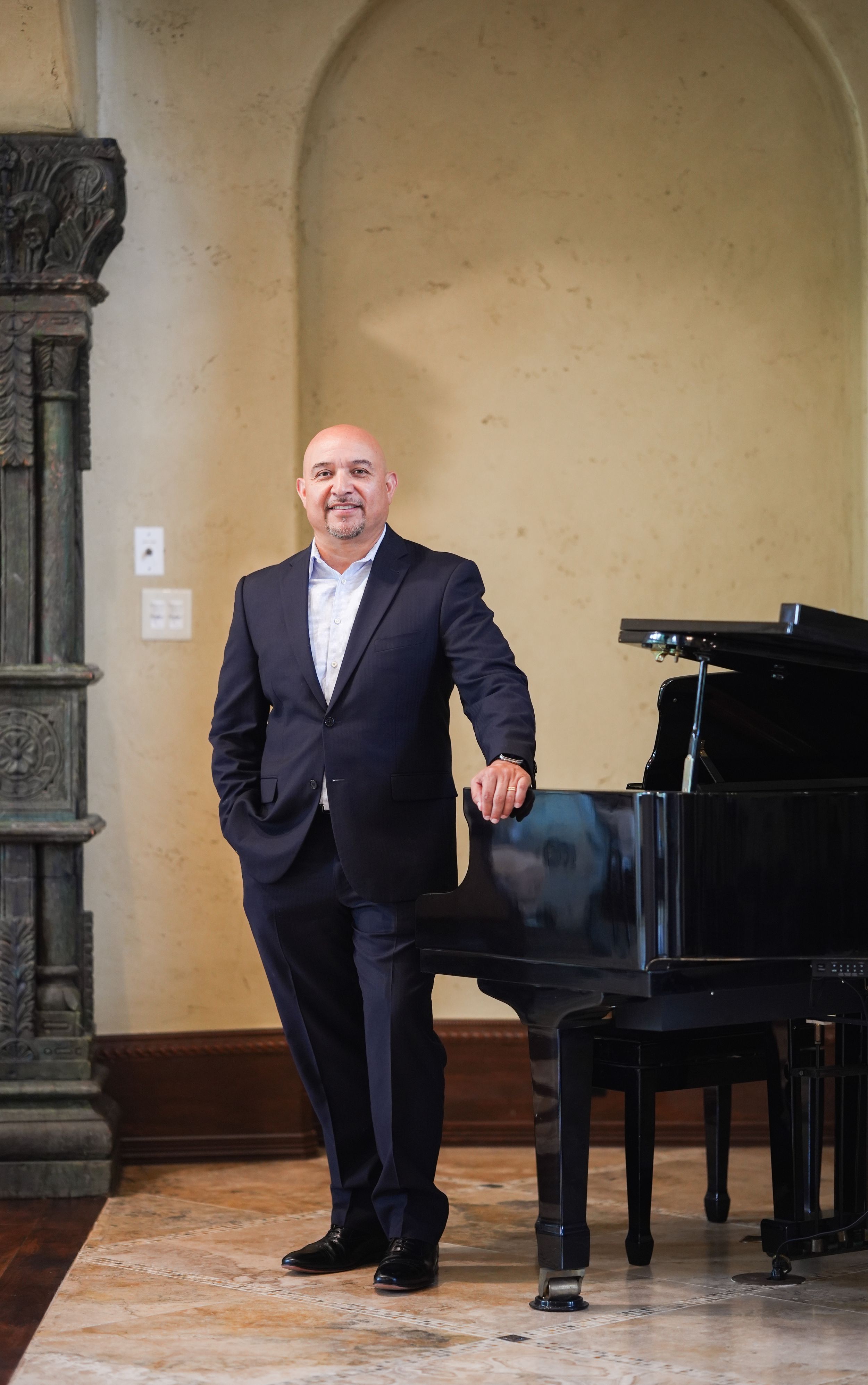 Man in dark suit leaning on grand piano in elegant room with arched walls and marble floors.
