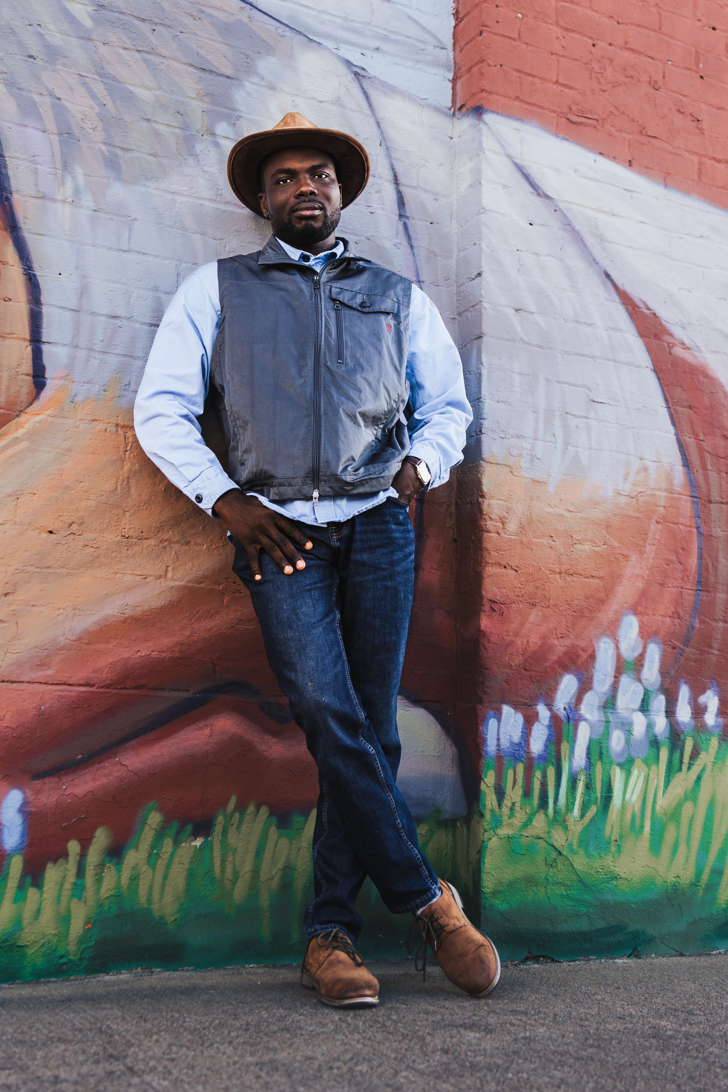 Man in hat and vest leans against colorful mural wall, wearing jeans and brown shoes in urban setting.