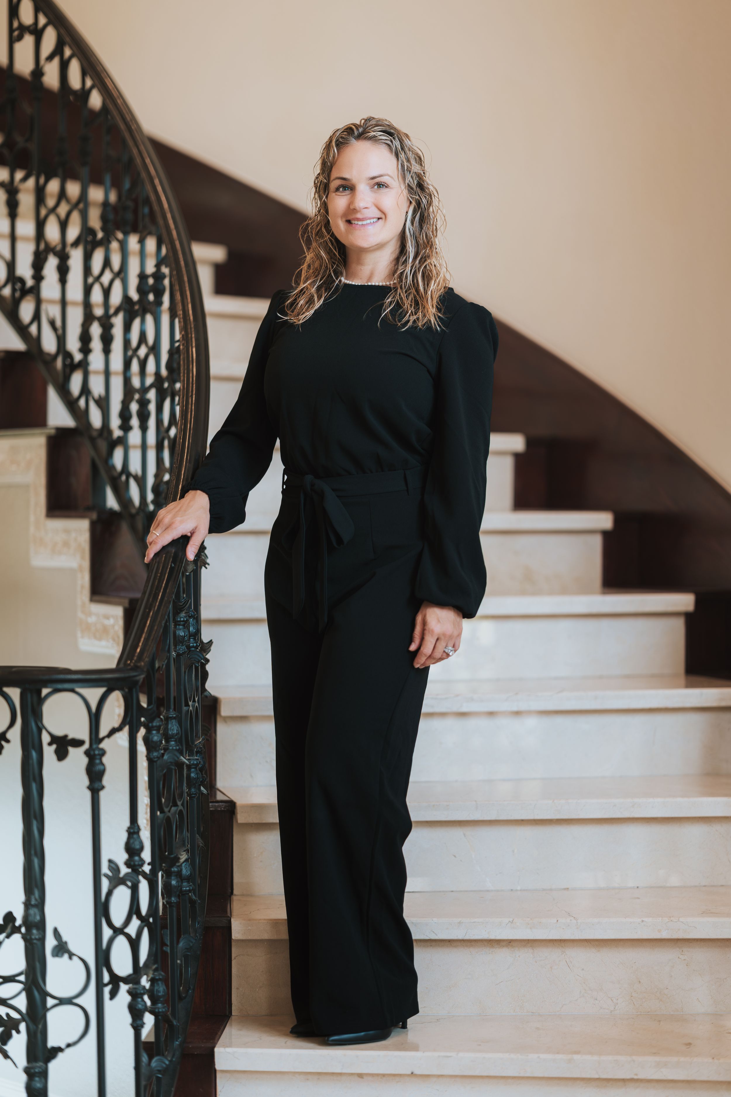 Woman in black jumpsuit smiling on elegant marble staircase with ornate iron railing.