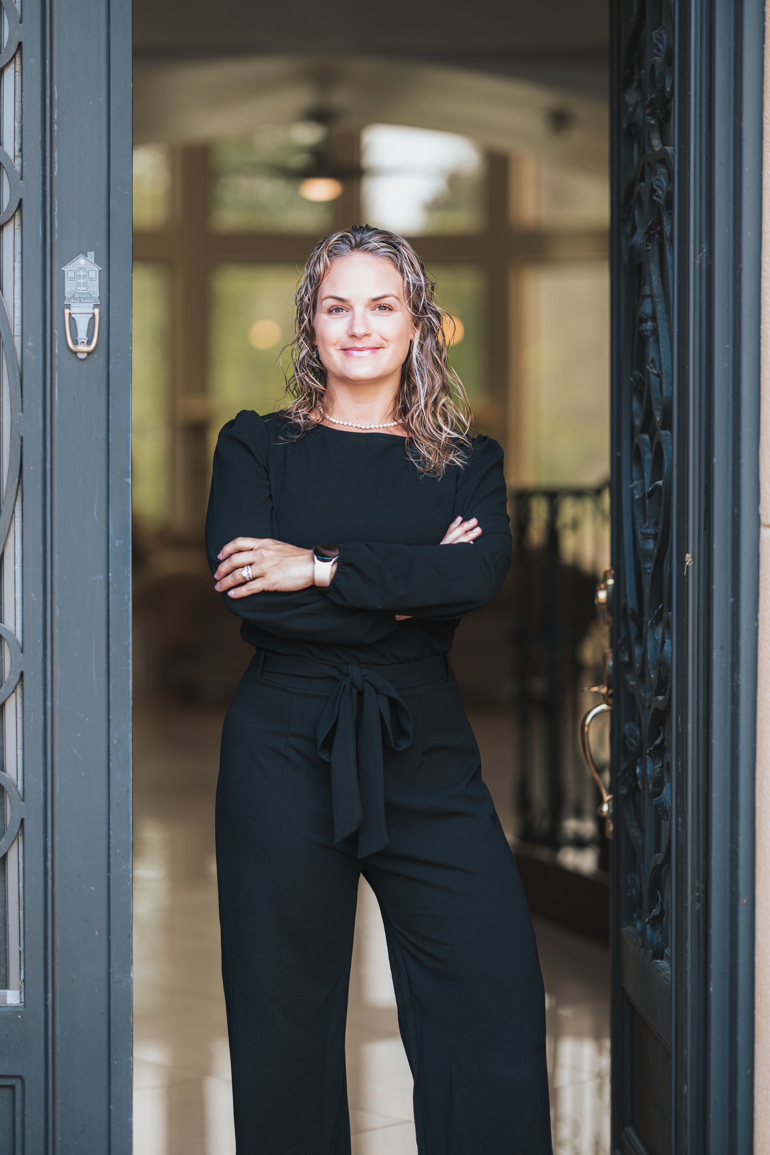 Professional woman in black jumpsuit stands confidently with arms crossed in elegant doorway, smiling warmly.
