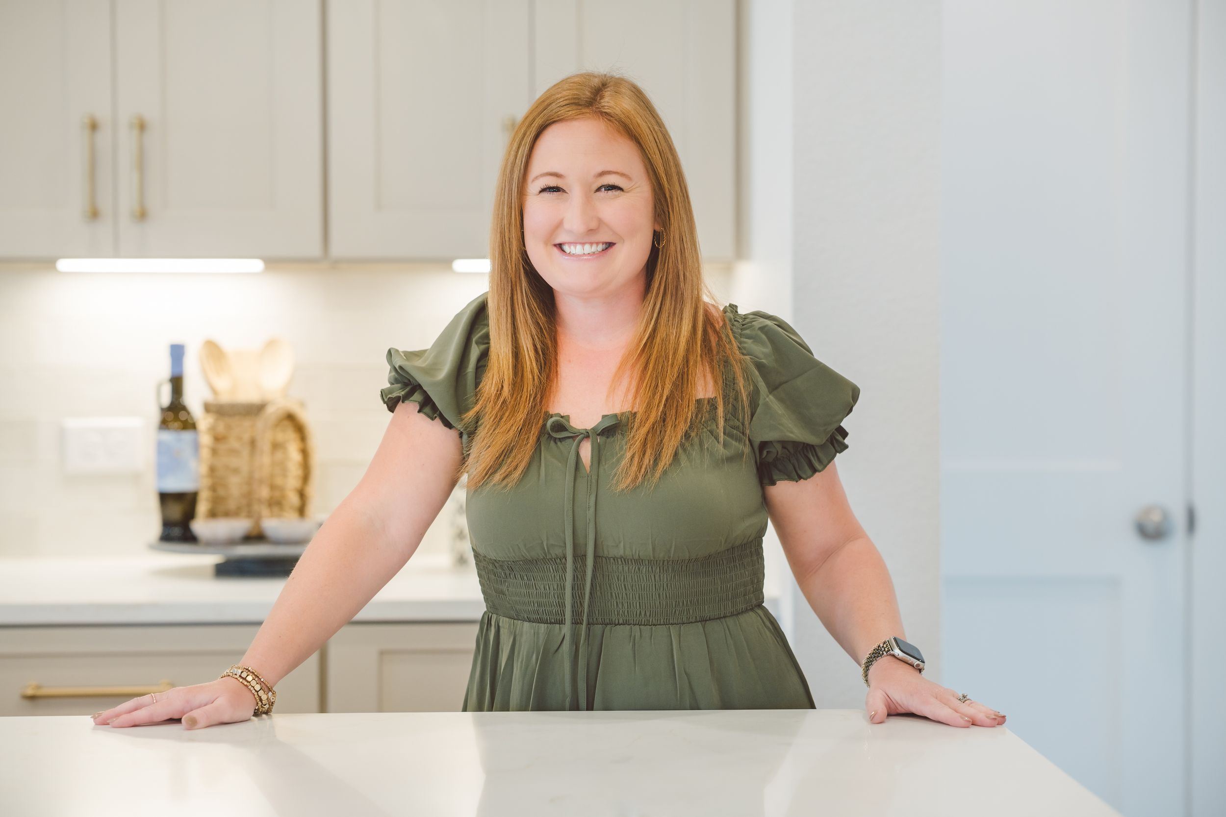 A woman in an olive green dress stands confidently in a bright modern kitchen with white cabinets and countertops.