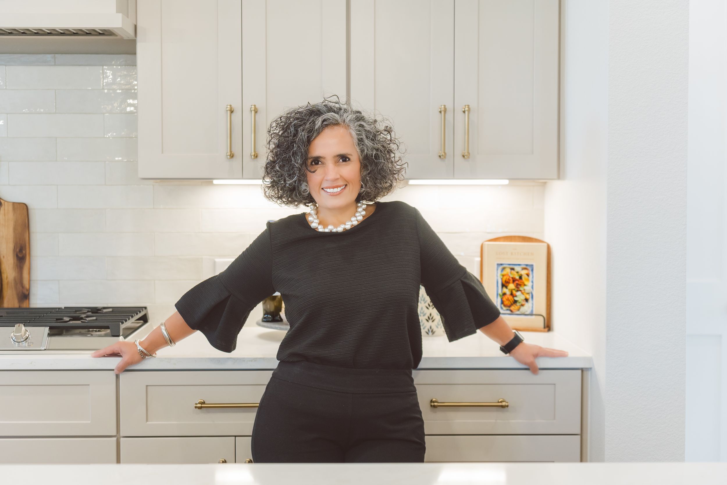 A person in black clothing poses in front of a white modern kitchen with light wood cabinets and stainless appliances.