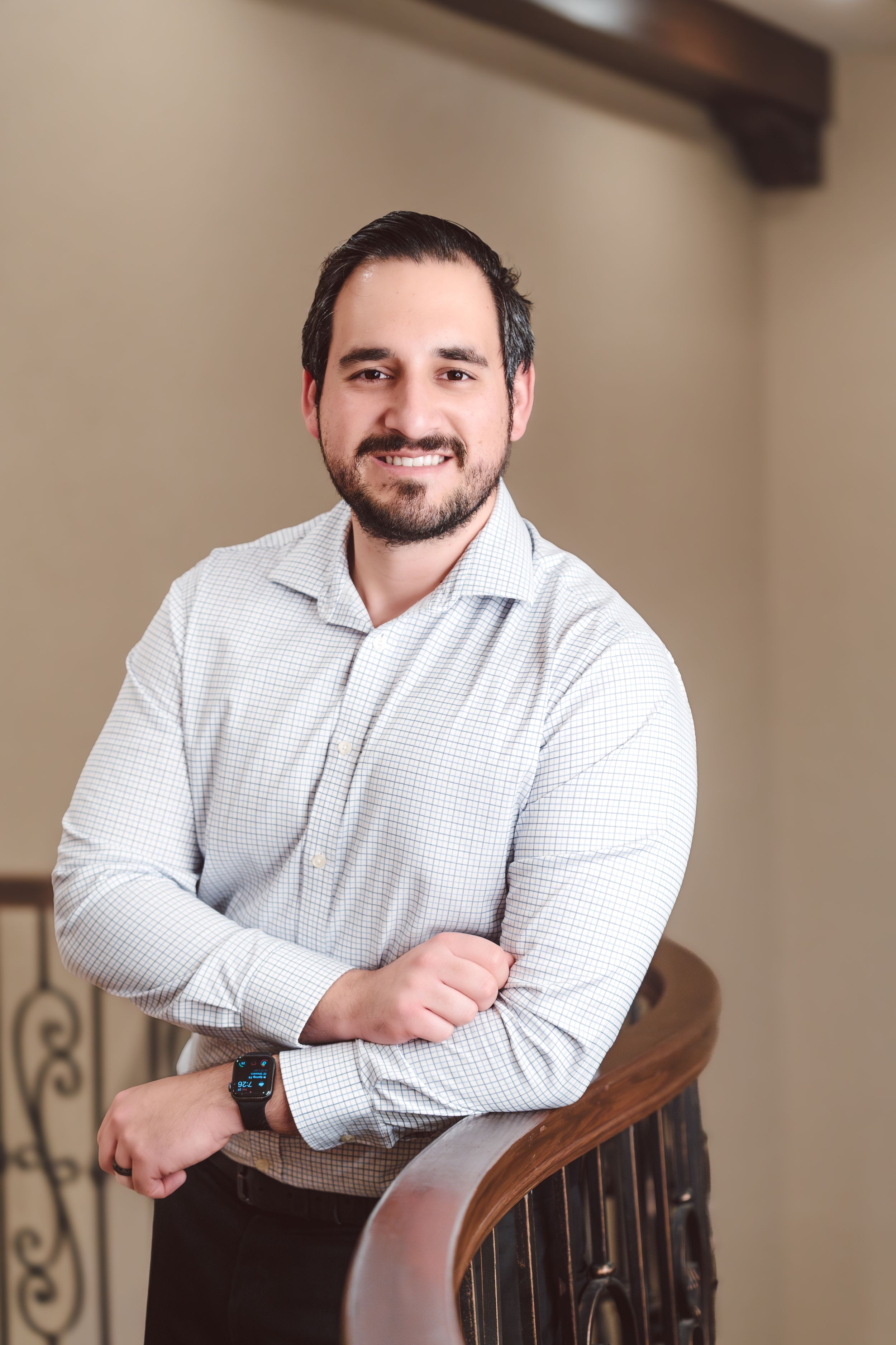 Professional headshot of someone in a white button-down shirt smiling while leaning on a wooden chair.