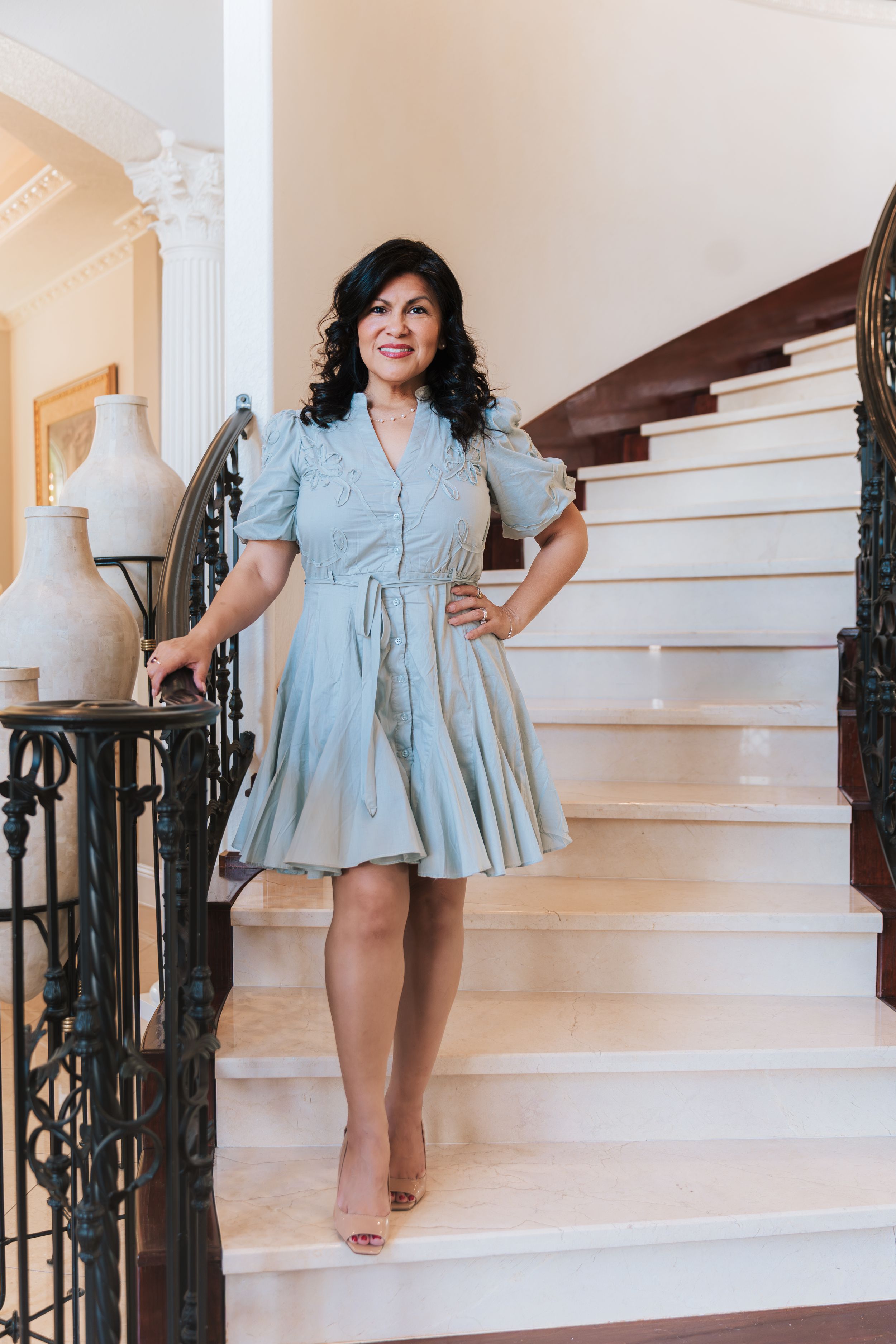 Woman in light blue dress posing on elegant staircase with wrought iron railing in a grand home.