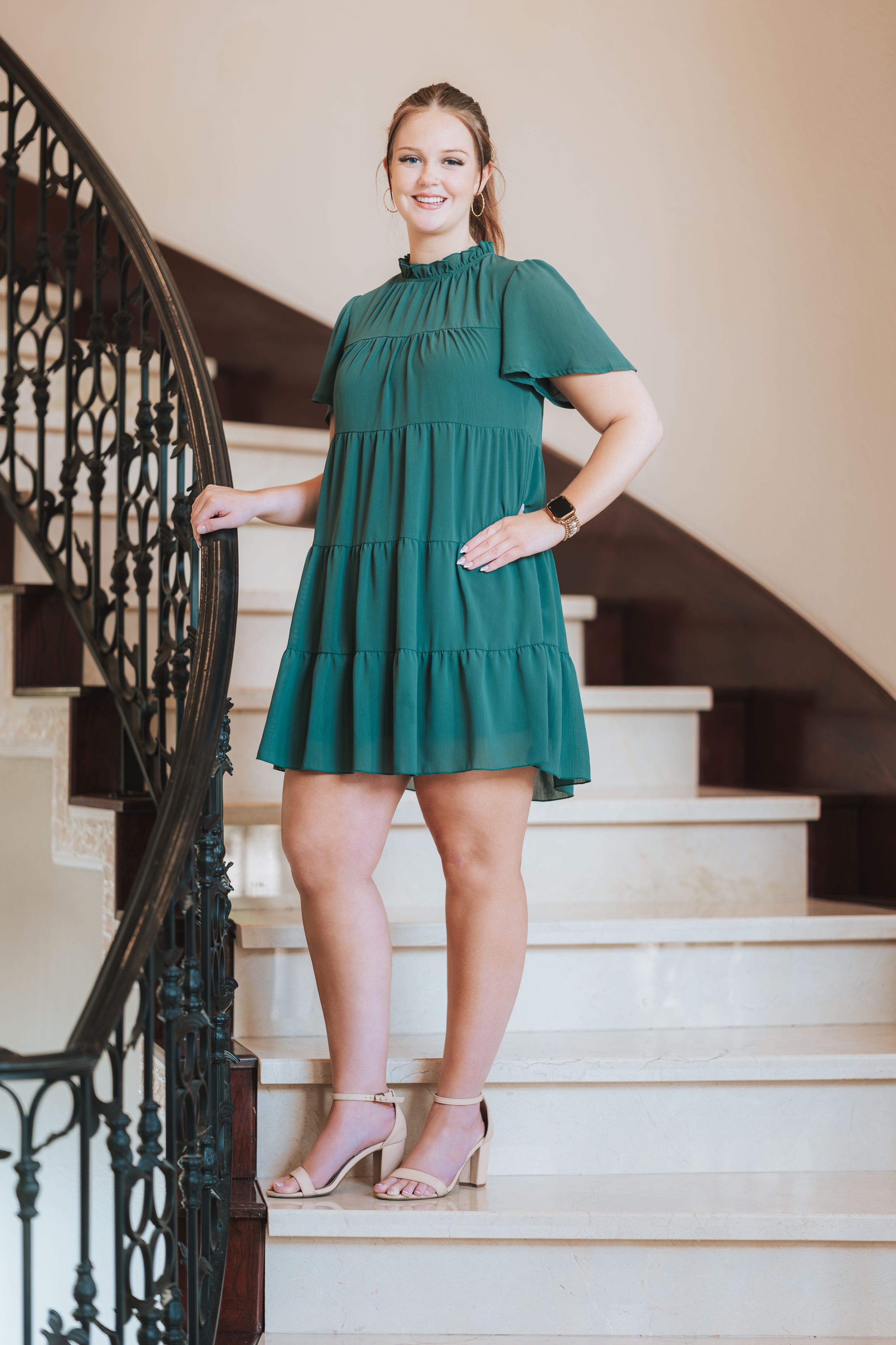 Woman in green tiered mini dress with flutter sleeves posing on elegant staircase with wrought iron railing.